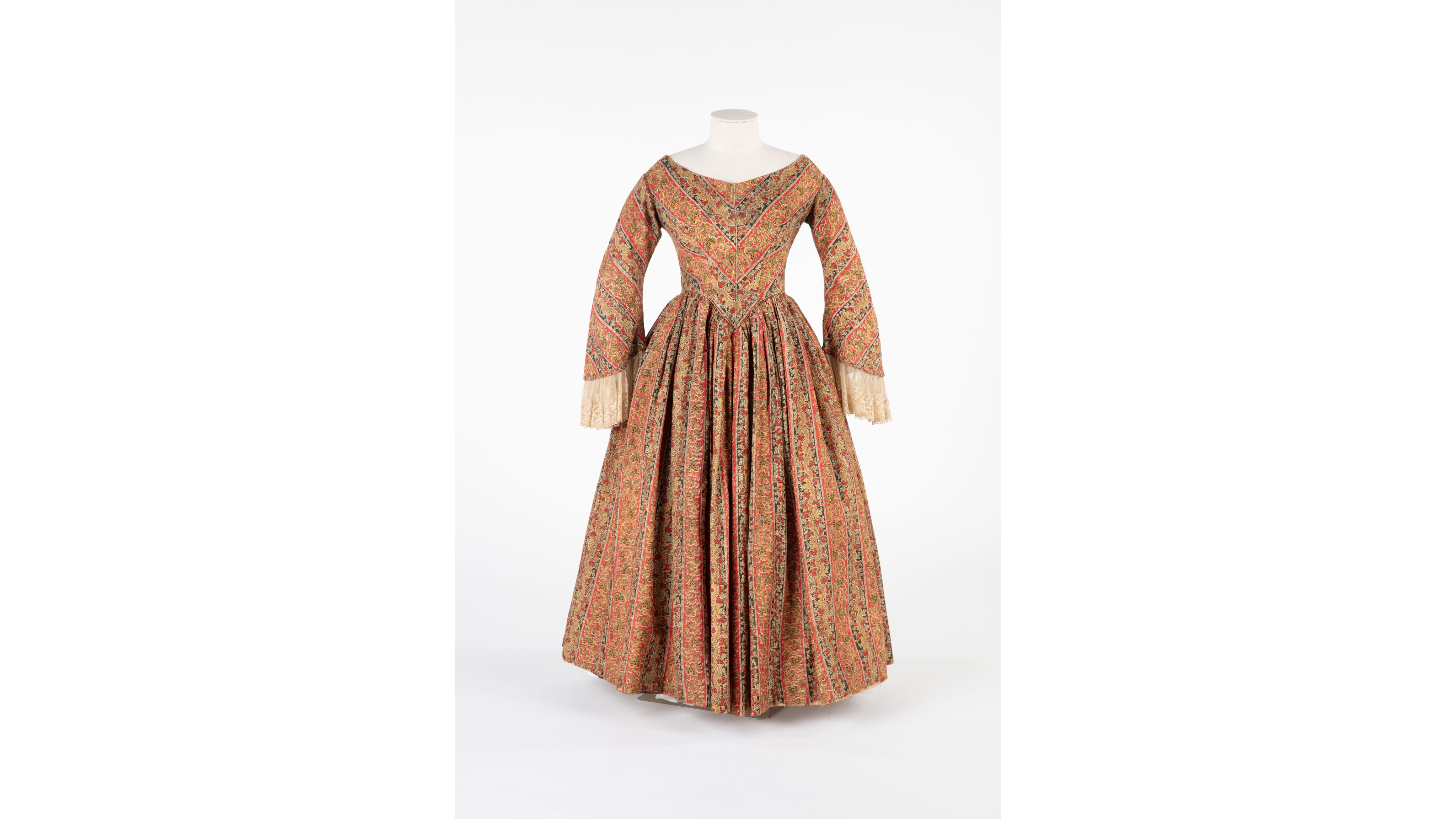 Copper-toned dress of fine delaine wool/cotton paisley with red and black striped and over-printed designs. The fitted, boned bodice features a wide V-neckline, green silk piping, and back fastening. Altered ¾ sleeves are wide at the forearm with decorated net flounces. The full skirt, lined and gathered at the waist, falls to a straight hem.
