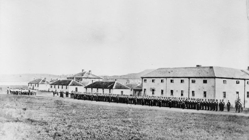 The image shows a group of men standing in front of a building with windows, surrounded by grass and hills in the background, with a clear sky above. 